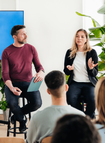 The image shows two people standing in front of a class or lecture. The woman on the right is talking to the group. The man on the left is in front of a screen and is holding a tablet. 